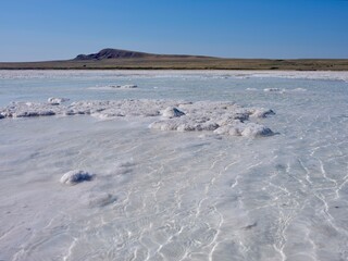 Shallow clear water over white salt crusts at Lake Baskunchak, a saline lake in Astrakhan Oblast, Russia. Rippled patterns and mineral deposits, Bogdo Mountain on the horizon, blue sky, no people.