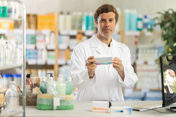 Fototapeta premium Male pharmacist stands behind the counter in a pharmacy, works at a computer and holds a package of vitamins and medicines. Pharmacy warehouse employee checks the availability of goods