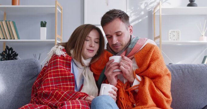 Married and loving couple embracing and staying warm under a blanket while drinking hot tea at home during cold winter weather. Representing comfort, love, and togetherness in domestic environment.