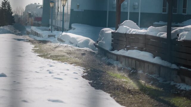 soft winter daylight illuminates snow covered street walkway alongside building benches and lamp posts melting snow reveals patch of grass with rising steam wisps creating serene urban winter scene
