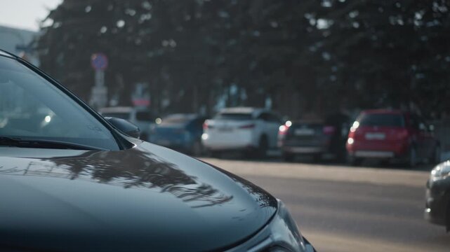 side view of cars parked along both sides of busy urban street with vehicles driving past streetlights and building facades in clear daylight, asphalt pavement, winter season with light snow remnants
