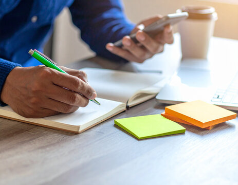 A person writes in a notebook with a pen while holding a smartphone, with a coffee cup, laptop, and sticky notes nearby on a desk
