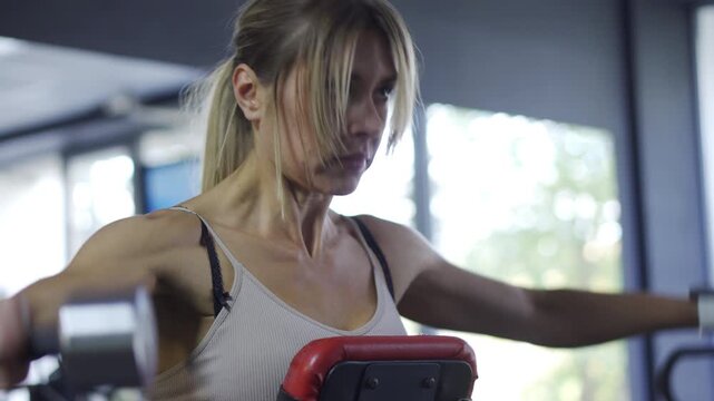 Shoulder Workout: Strong blonde woman doing lateral dumbbell raises on a bench in the fitness club