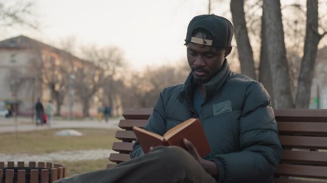 Young man in winter jacket sits on wooden park bench reading hardcover book with deep focus, legs crossed, cap backward, evening light, blurred people passing in background