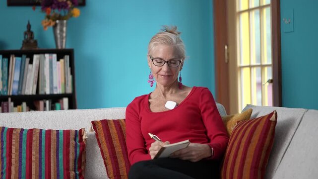 Mature woman relaxes on the couch in a bright living room, wearing a heart sensor to monitor atrial fibrillation and maintain her health.