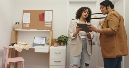 Diverse coworkers entering fashion studio, tapping tablet and lifting dress for comparing fit - Powered by Adobe