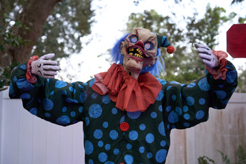 A creepy Halloween clown with spiral eyes and a wide grin stands with open arms. Blue hair, red nose, yellow teeth, and a green polka-dot suit with orange ruffle and buttons fill the outdoor scene.