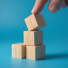 Finger knocking over a small tower of three blank wooden cubes, blocks, pushing and destabilizing a built structure, loss of balance simple abstract concept. Object closeup, detail, blue background