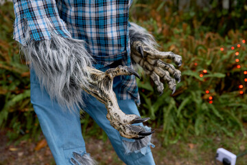 Close-up of a werewolf's clawed hands reaching forward with sharp black nails. Hairy gray fur, plaid sleeves, and torn jeans create a spooky outdoor scene.