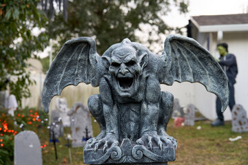 A winged stone gargoyle crouches on a pedestal in a Halloween graveyard. Tombstones, lights, and a spooky yard create a haunted outdoor scene.