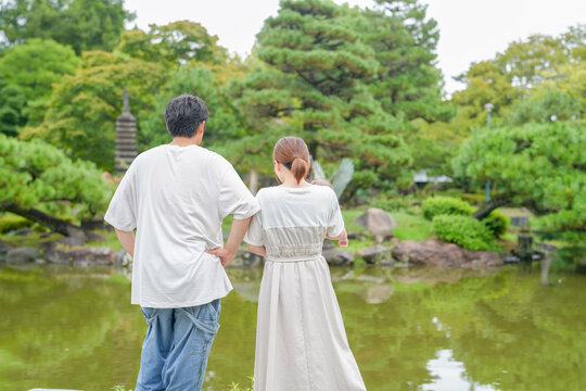 A man and woman are standing by a pond in a park. The man is wearing a white shirt and jeans, while the woman is wearing a white dress. They are both looking at the water, and the scene has a peaceful - Powered by Adobe