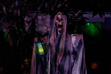 A hooded skull figure in a tattered gray cloak stands at night with a glowing blue-green light. Spooky Halloween yard decoration with dark background and eerie mood.