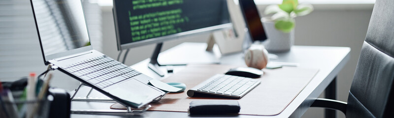 Header of modern office workspace featuring desktop computer with code on screen, wireless keyboard, mouse, laptop on stand, smartphone and accessories arranged on desk in bright environment
