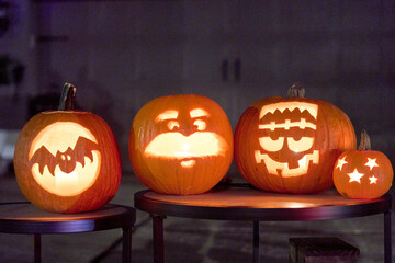Four carved pumpkins with bat, cartoon face, stitched monster, and star cutouts glow on round tables at night. Warm orange candlelight creates a spooky, festive Halloween display in a suburban