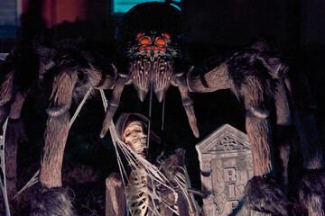 A huge hairy spider with red eyes looms over a skeleton beside an RIP tombstone. Creepy Halloween yard decoration with webs, fangs, and eerie lighting on the lawn.