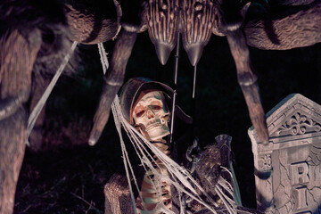 A Halloween skeleton sits beneath a giant spider with fangs, webs, and red eyes beside an RIP tombstone. Spooky yard decoration with dark lighting on a suburban lawn.