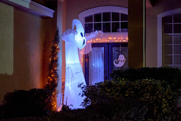 A tall white ghost inflatable glows beside the front door with orange string lights at night. Halloween yard decorations create a spooky, festive entrance to a suburban home.