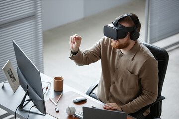 Caucasian young adult man sitting at desk using virtual reality headset and interacting with digital interface, computer monitor and office supplies visible on workspace