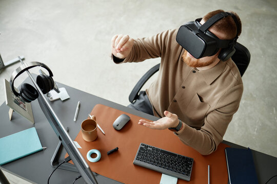Caucasian young adult man using virtual reality headset while sitting at desk, interacting with digital objects in air, working with computer and office accessories visible on workspace