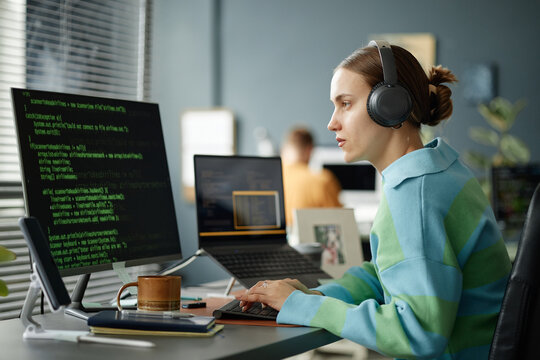 Caucasian young adult woman wearing headphones working on computer with code displayed on monitor, sitting at desk in modern office environment, focusing on programming tasks