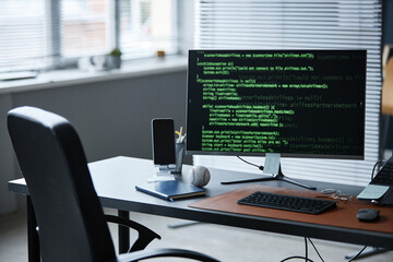Modern office workspace featuring desktop monitor displaying programming code, wireless keyboard, smartphone on stand, baseball and eyeglasses resting on desk, window blinds in background