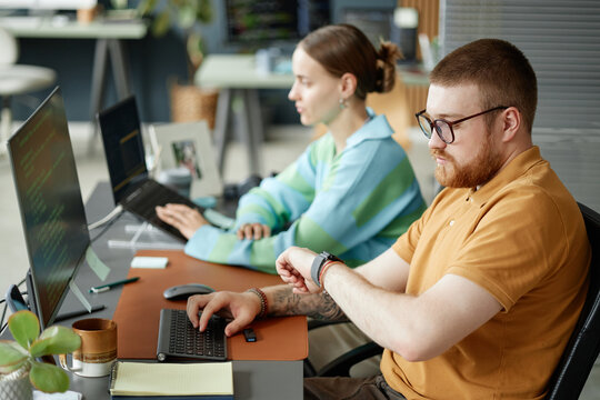Caucasian young adult man checking smartwatch while working at computer next to Caucasian young adult woman typing on laptop in modern office setting, both focused on tasks