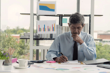 Focused senior asian businessman in an office intently reviewing and writing on document surrounded...