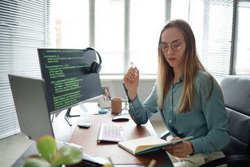 Caucasian young adult woman sitting at desk working on computer with code on screen, holding pen and notebook, wearing glasses, focused on programming in modern office environment