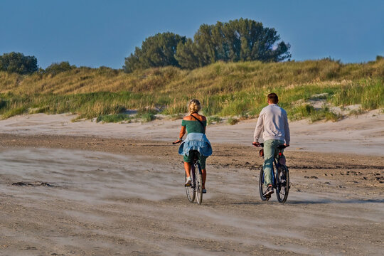 A couple riding bicycles on a sandy beach near grassy dunes during a sunny day. Active lifestyle and leisure at the seaside.