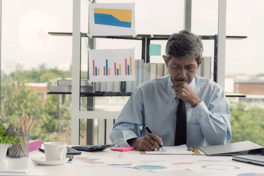 Focused senior asian businessman in an office intently reviewing and writing on document surrounded by chart and graph indicating deep concentration and financial analysis.