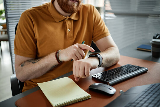Caucasian young adult man sitting at desk checking smartwatch while holding pen near notepad and computer keyboard, focusing on time management and productivity in office setting - Powered by Adobe