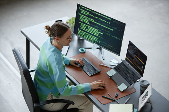 Caucasian young adult woman working at desk using desktop computer and laptop with programming code on screens, holding mouse and typing on keyboard, surrounded by office supplies - Powered by Adobe