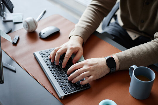 Young adult man typing on wireless keyboard at modern desk, hands visible with smartwatch, coffee mug and baseball on workspace, working in office environment - Powered by Adobe