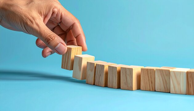 A hand halts a curved line of falling dominoes&mdash;placing one upright block to interrupt the chain reaction on a blue surface. A metaphor for intervention and the choreography of consequence through deci