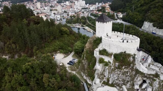 Medieval cliff-top fortress overlooking river and red-roofed town in Uzice, Serbia, with winding road. g.