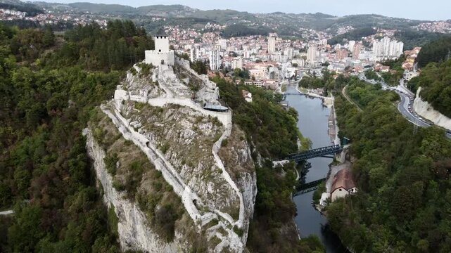 Medieval cliff-top fortress overlooking river and red-roofed town in Uzice, Serbia, with winding road. g.