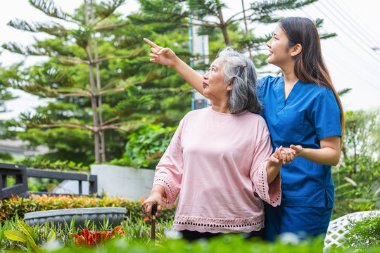 Happy senior woman walking with a young nurse in a lush green outdoor area, Young Asian caregiver assisting a senior woman with a walking stick in a park