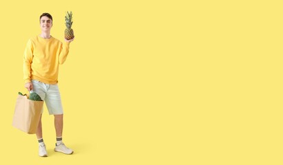 Young man holding pineapple and paper shopping bag on yellow background with space for text