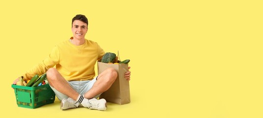 Young man with full shopping basket and paper bag sitting on yellow background with space for text