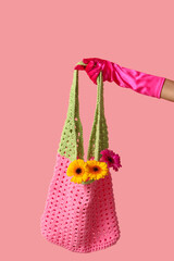 Female hand in laced glove holding crocheted bag with gerbera flowers on pink background