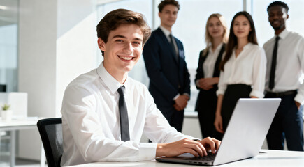 A young professional man is using a laptop in an office with a diverse team standing behind him, indicating a collaborative work environment.