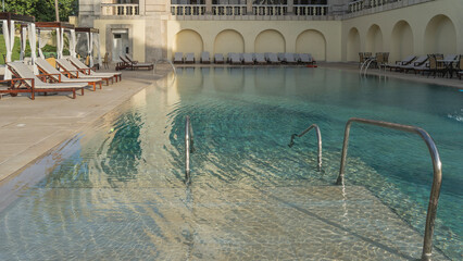 Swimming resort pool. Clear water, turquoise tiled bottom, metal handrails of stairs. Rows of deck chairs and awnings with curtains around the perimeter. Cuba. Havana. Hotel