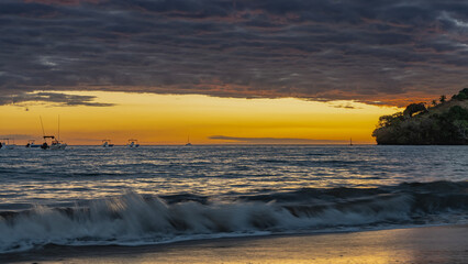 A bright sunset on a tropical island. Ocean waves are foaming on a sandy beach. Blue clouds are highlighted in pink. The sky is orange on the horizon. Silhouettes of boats in the sea. Madagascar.