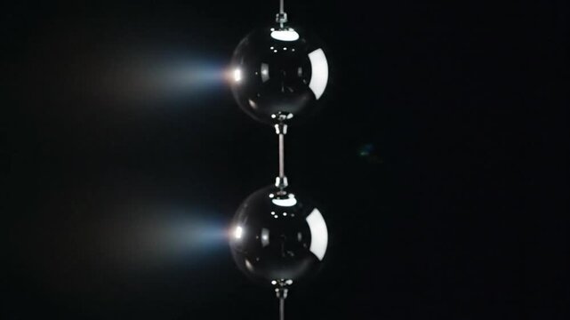 Close-up of two shiny metallic spheres hanging from a thread against a dark background with reflective light.