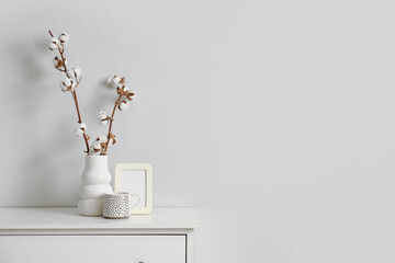 Fototapeta premium Photo frame, cup and vase with cotton flowers on chest of drawers near white wall in living room, closeup