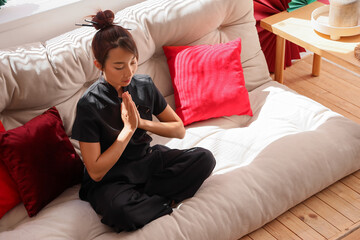 Obraz premium Young asian woman meditating in prayer pose on sofa, practicing mindfulness and relaxation at home, she is sitting cross-legged with her eyes closed and hands folded