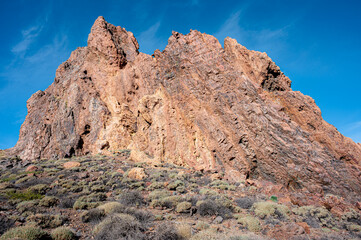 Fototapeta premium Low angle shot of a large orange volcanic rock rising from desert terrain, surrounded by sparse vegetation and native plants at Cañadas del Teide, Tenerife. 