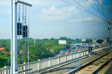 Fototapeta premium View from the Jakarta–Bandung high-speed train window showing elevated railway tracks above a busy toll road with green trees and a bright blue sky in the background.