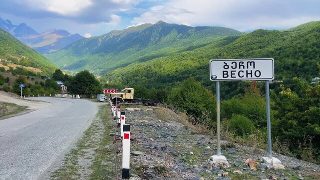 Mountain road leads into Becho village, Georgia, with a signpost in Georgian and English set against lush green hills and dramatic Caucasus peaks under a bright sky.