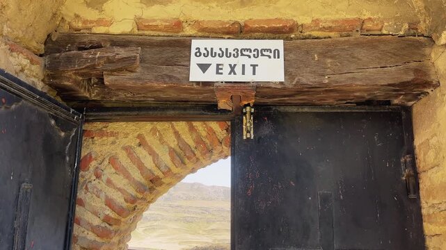 Exit doorway at Davit Gareja Monastery, Georgia, with a rustic wooden beam and bilingual sign. Historic architecture and cultural heritage in a scenic mountain setting.